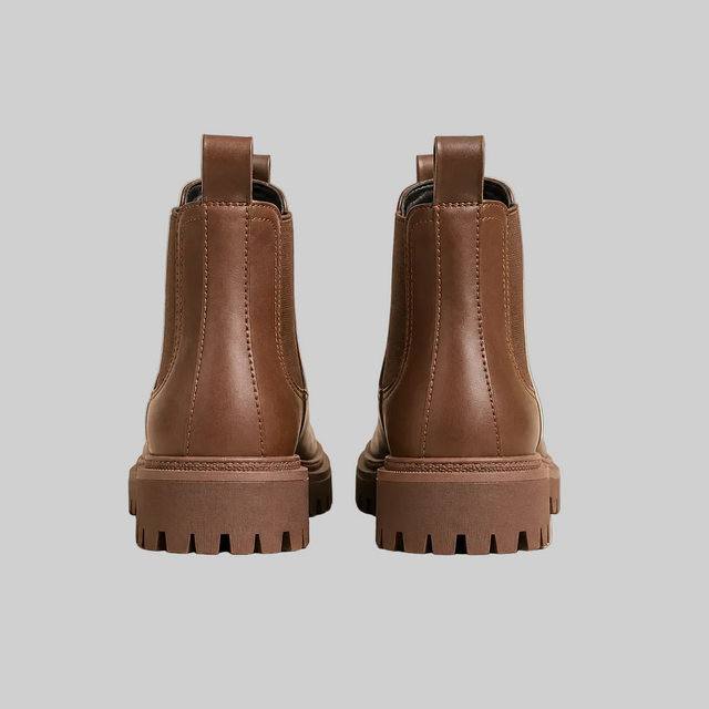 A pair of brown leather Chelsea boots with chunky lug soles, shown from the back on a light beige background.