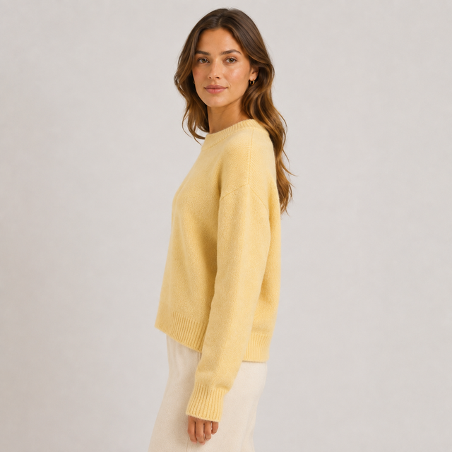 A woman wearing a pale yellow knit sweater and cream pants, photographed against a plain light gray studio background.
