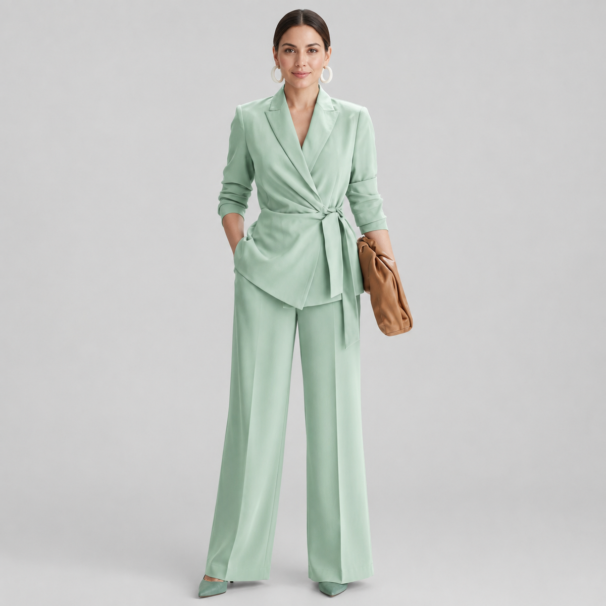 A woman models a sage green wrap blazer and matching wide-leg trousers set, styled with a tan clutch and matching green pointed shoes against a light gray studio background.