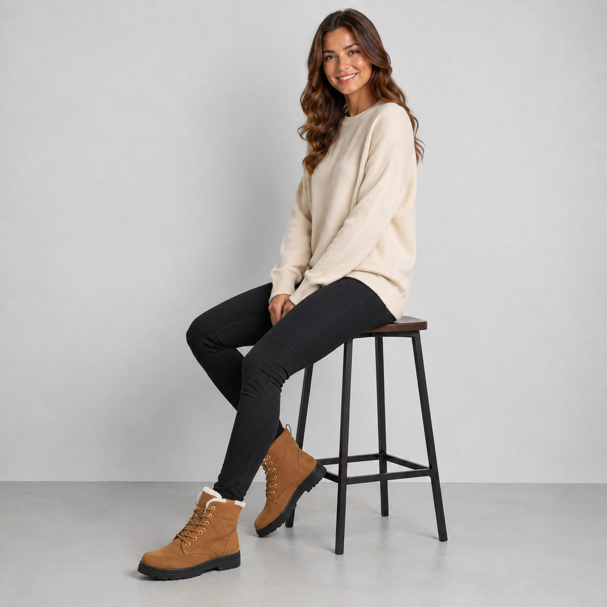 A woman sits on a black metal stool wearing a cream crewneck sweater, black skinny pants, and tan lace-up boots with white lining, against a light gray studio background.