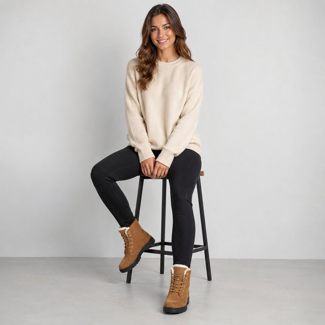 A woman sits on a black metal stool wearing a cream crewneck sweater, black skinny pants, and tan lace-up boots with white lining, set against a plain light gray studio background.