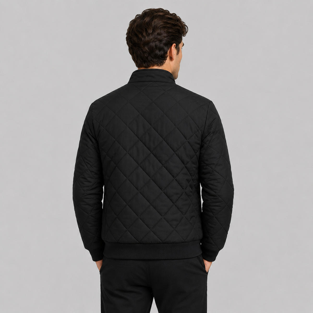 A man viewed from the back wearing a black diamond-quilted bomber-style jacket and black trousers, standing against a solid light gray studio background.