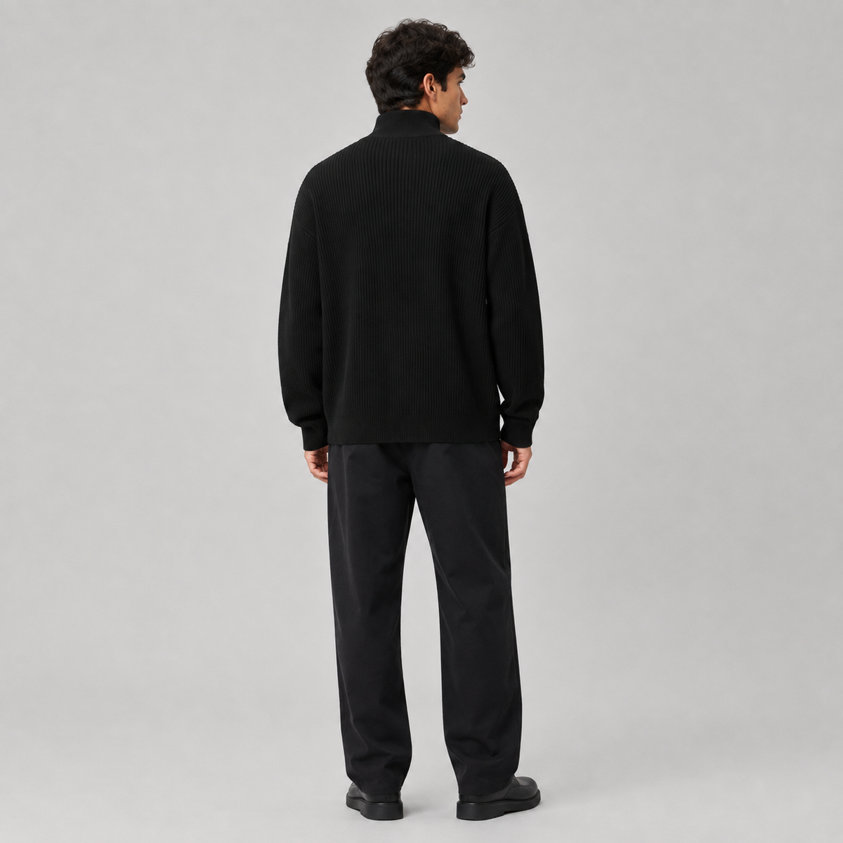 A man viewed from the back wearing a black ribbed mock-neck sweater and black trousers, standing against a light gray studio background.