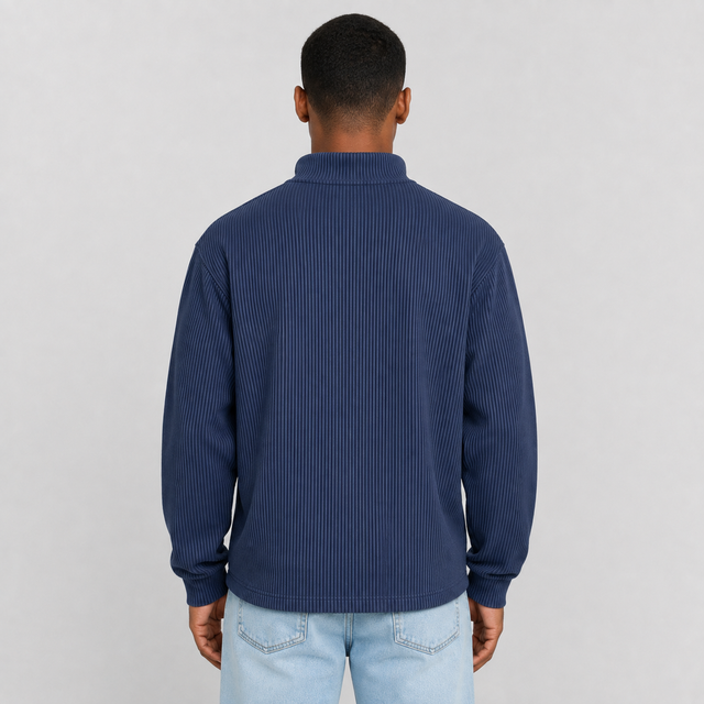 A man viewed from the back wearing a navy blue ribbed long-sleeve mock neck sweater and light wash jeans, set against a plain light gray studio background.
