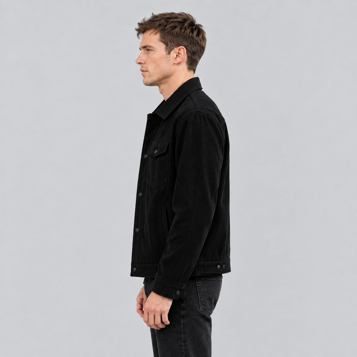 A man in profile wearing a black corduroy jacket and dark jeans, standing against a light gray studio background.