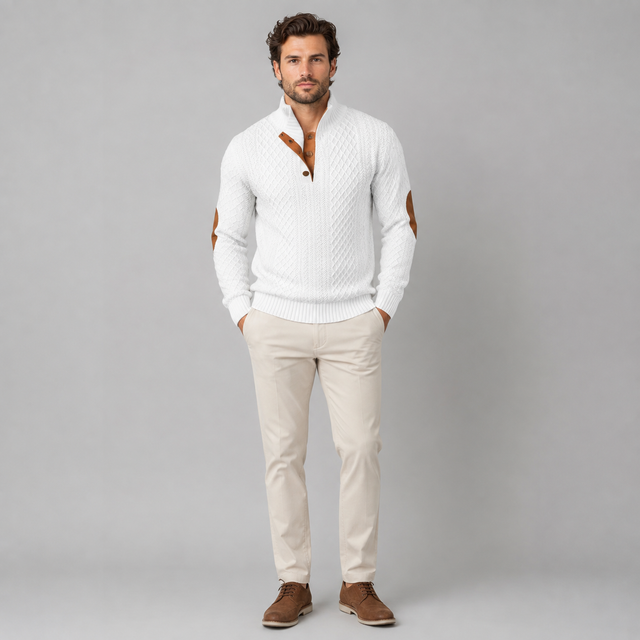 A man modeling a white cable-knit mock-neck sweater with brown leather elbow patches and collar trim, paired with beige trousers and brown shoes, against a solid gray studio background.