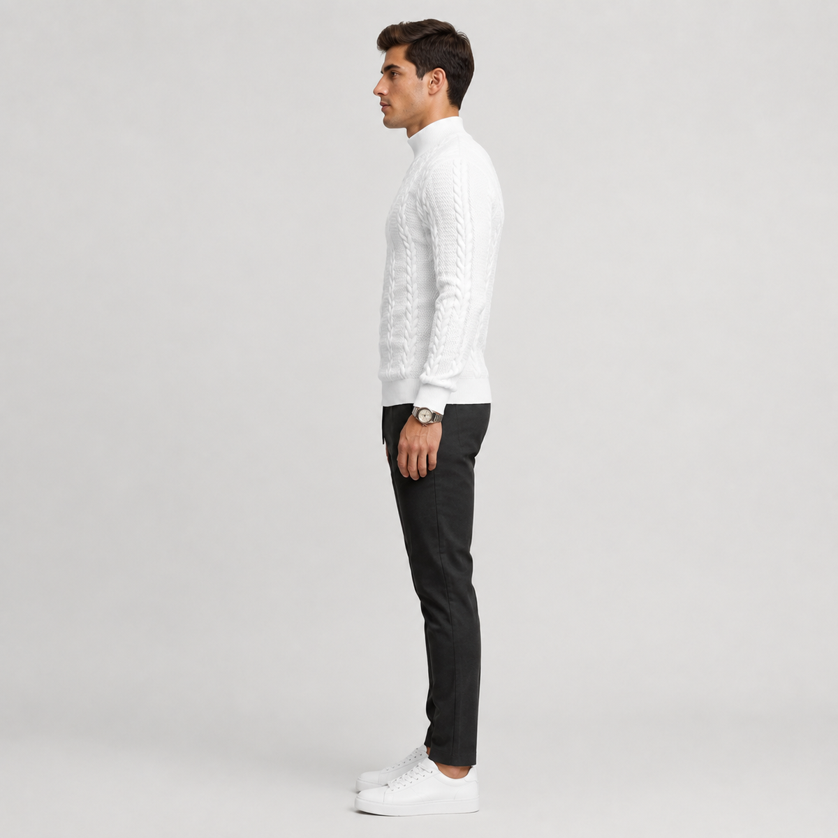 A man in profile wearing a white cable-knit mock-neck sweater, black trousers, and white sneakers, standing against a light gray studio background.