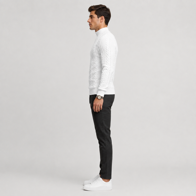 A man in profile wearing a white cable-knit mock-neck sweater, black trousers, and white sneakers, standing against a light gray studio background.